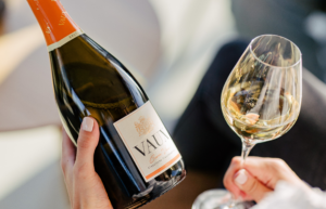 Jemand hält eine VAUX Cuvee Flasche in der linken Hand und und in der rechten Hand ein gefülltes VAUX Sektglas.