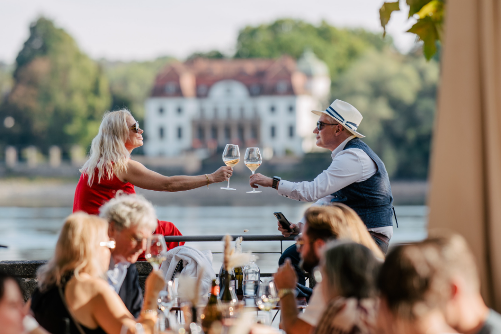 Im Vordergrund sitzen unscharf viele Menschen im Sektgarten. Im Hintergrund und vor dem Rhein ein Paar das anstoßt
